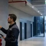 Technician inspecting a failing building safety system in Germany, showing fire alarm warnings and asset management compliance risks.